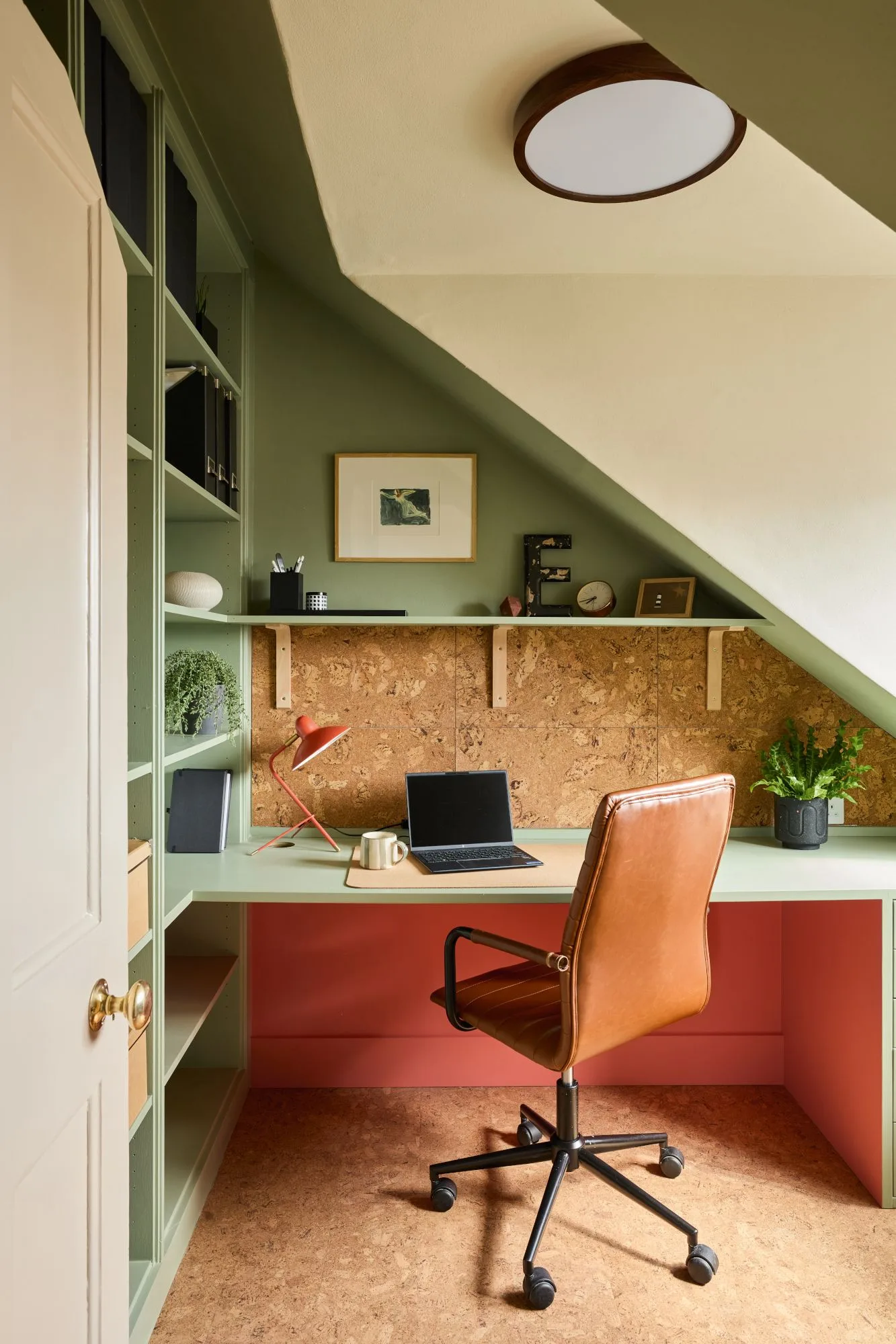 Compact home office under sloping ceiling with green shelves and desk, textured tan wall backing, coral lower panel, tan rolling chair, red lamp and small plant.