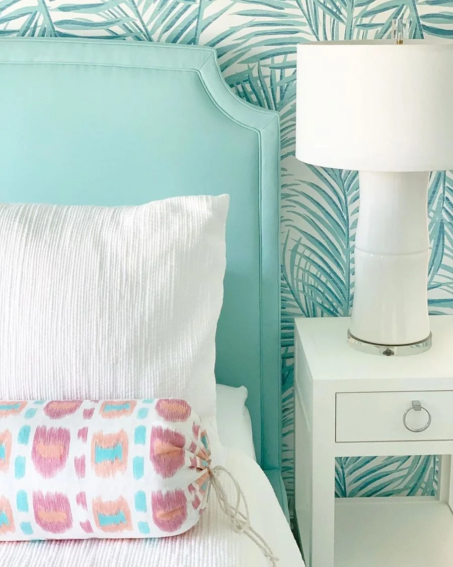 Turquoise palm-frond wallpaper connects with a pale turquoise upholstered headboard beside a white bedside chest and tall white lamp
