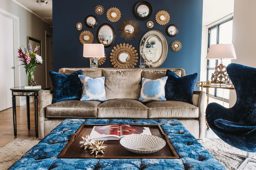A blue wingback chair stands beside a taupe sofa and patterned blue ottoman below a mirror collection