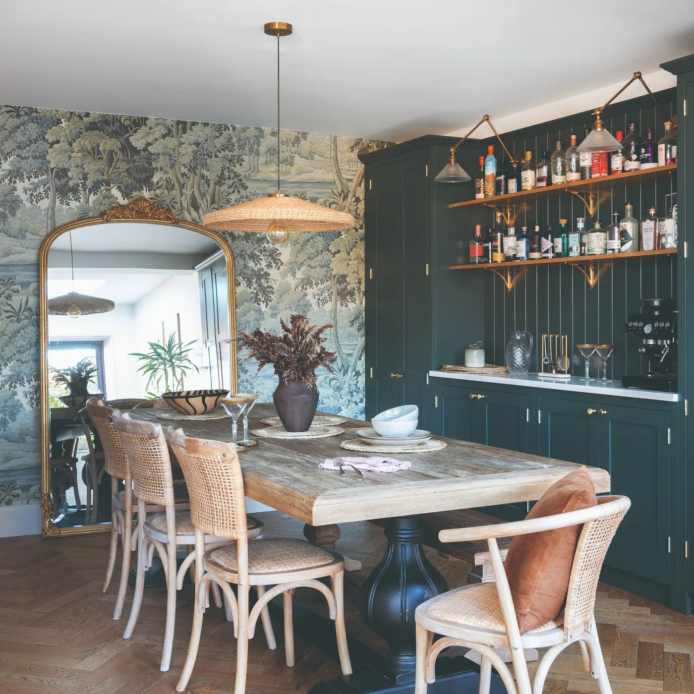 Broad woven pendant above a long wood dining table with woven chairs and botanical wallpaper beyond