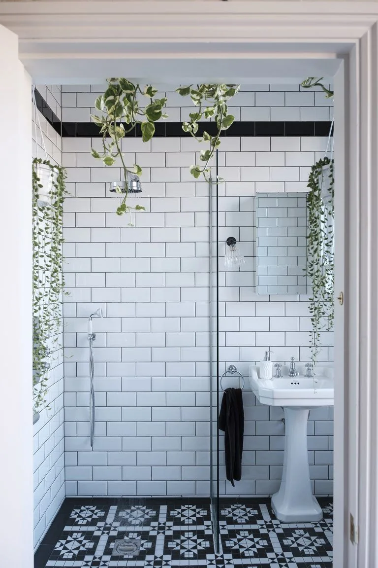 White subway tiles with dark grout and a black horizontal border sit above a pedestal basin and black-and-white patterned floor beside hanging plants