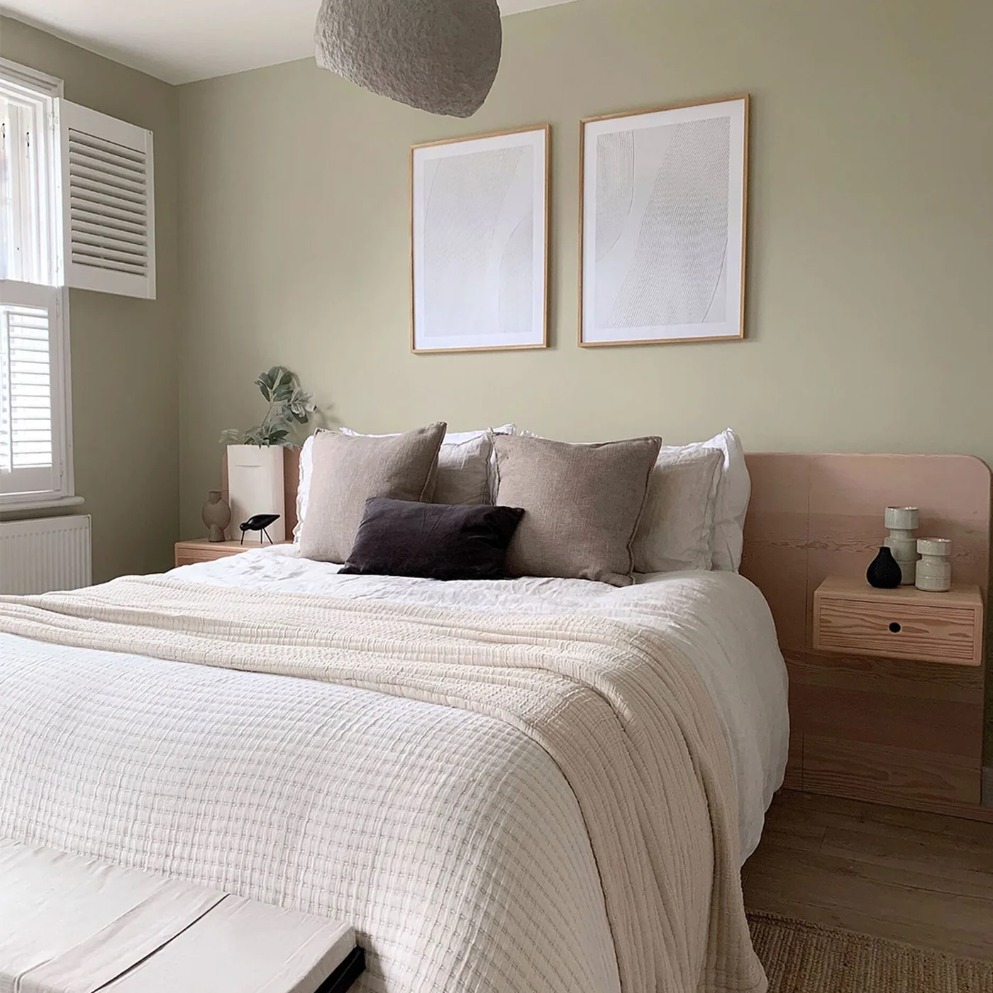 Muted green walls surround a pale upholstered headboard, light bedding, wooden bedside drawers and two framed pale pictures