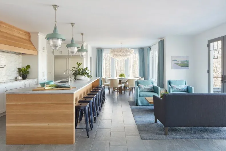 Open coastal room with wooden kitchen island, dark stools, blue living chairs and separate dining table near pale blue curtains.