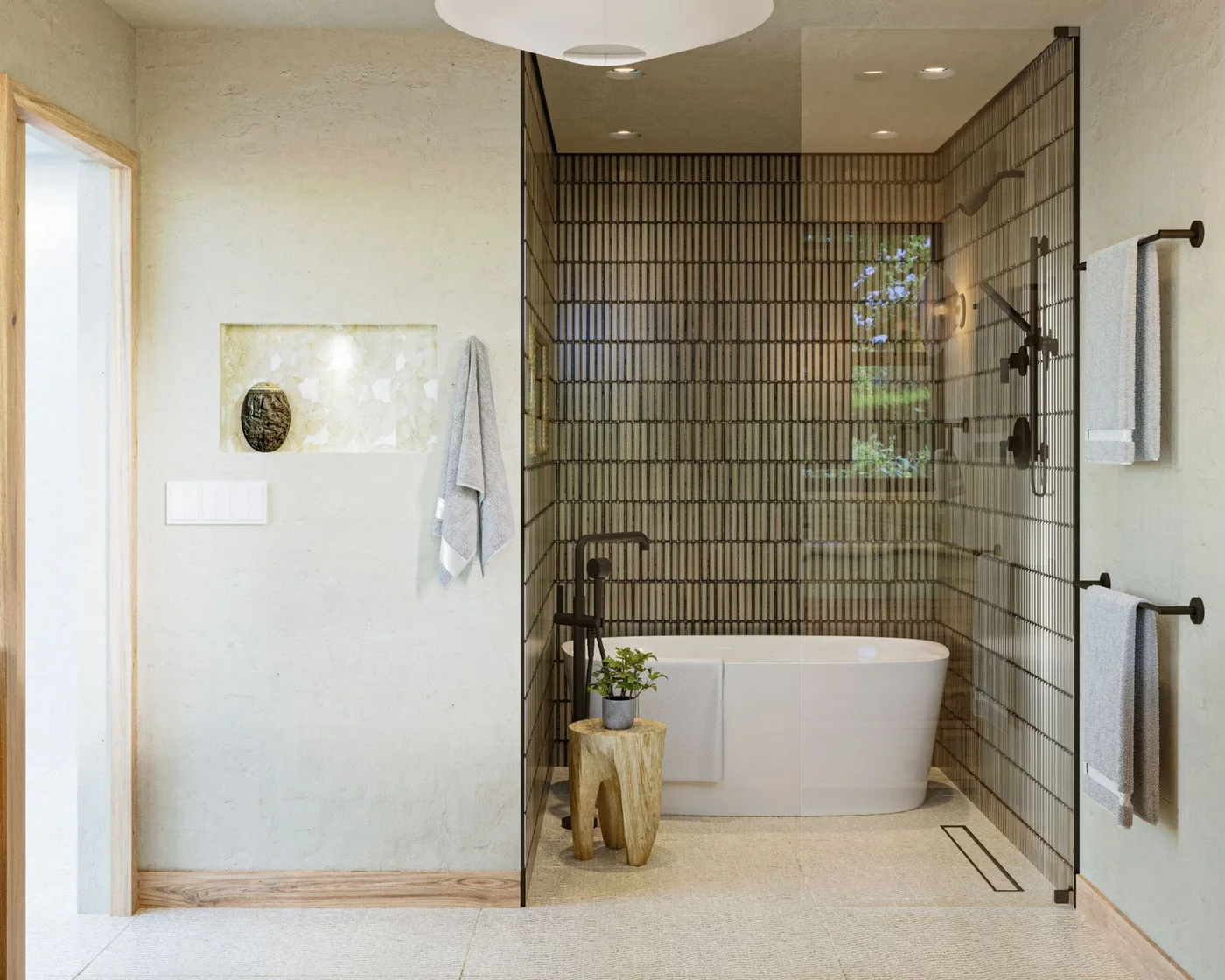 A freestanding white bath sits within a glass shower enclosure against narrow dark tiles beside pale walls, towel rails and a small wooden stool