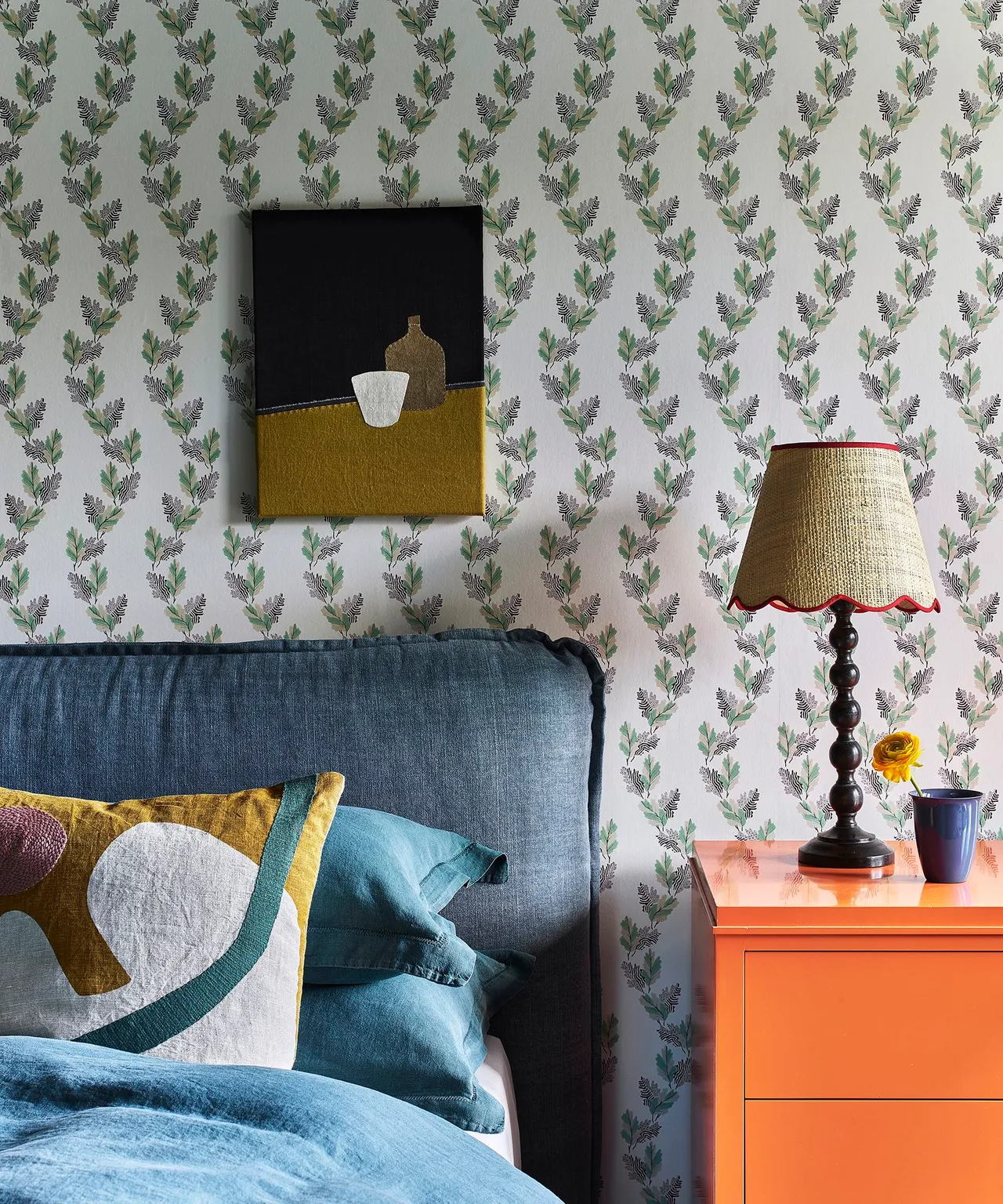 Bedroom with dark blue headboard and bedding, small botanical wall pattern, orange bedside drawers, turned dark lamp base and framed abstract artwork.