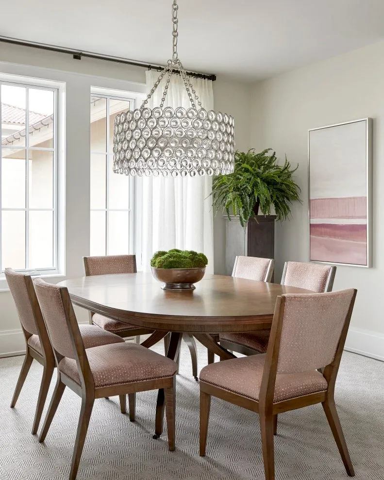 Pale pink upholstered chairs around a dark curved wooden table beneath a perforated ring-shaped chandelier.