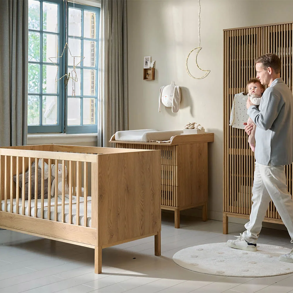 A parent holds a baby beside a wooden crib, slatted wardrobe and matching changing cabinet in a pale nursery