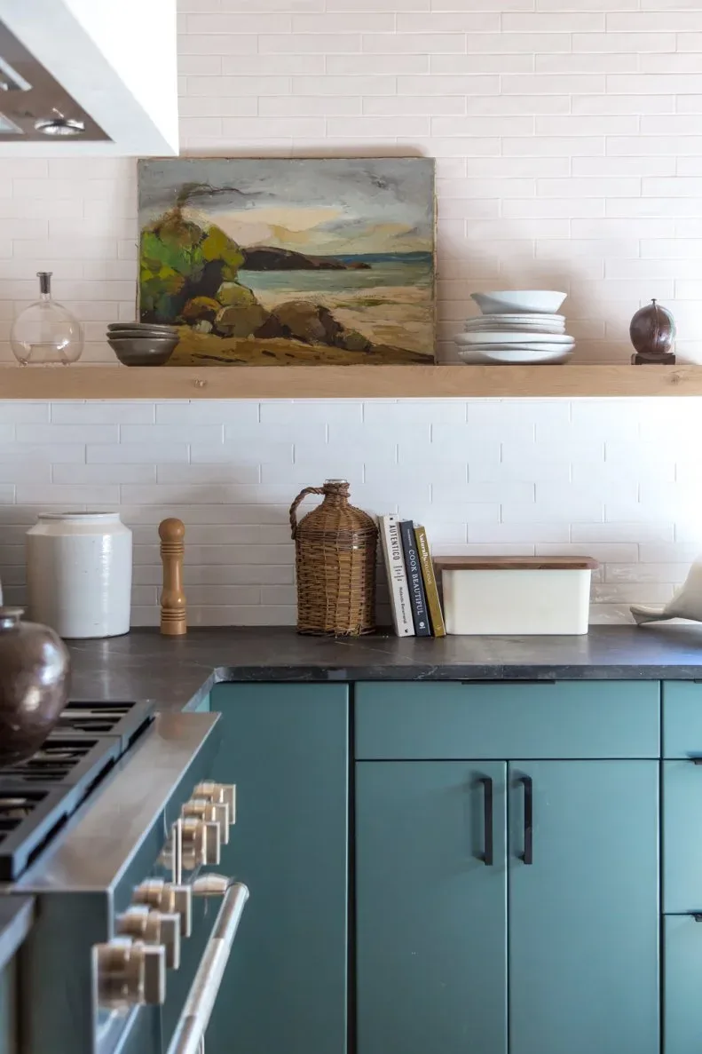 Teal lower cabinets beneath white tile, a wood shelf with a landscape painting and stacked bowls.