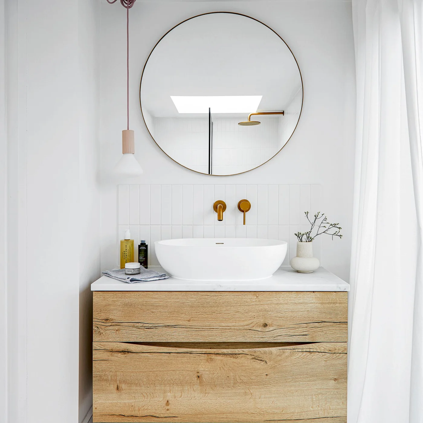 A wood-fronted vanity with a white bowl basin sits below a round mirror beside white walls, gold-colored wall taps and a small pendant