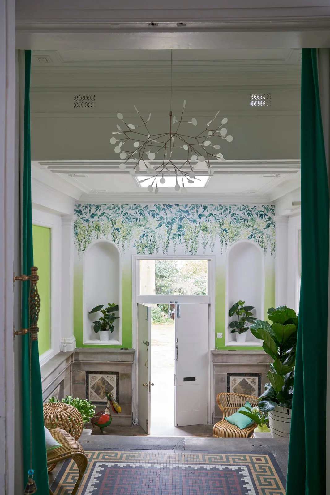 A leafy mural runs above a white front door between lime-green niches, woven chairs, plants and a patterned mosaic floor