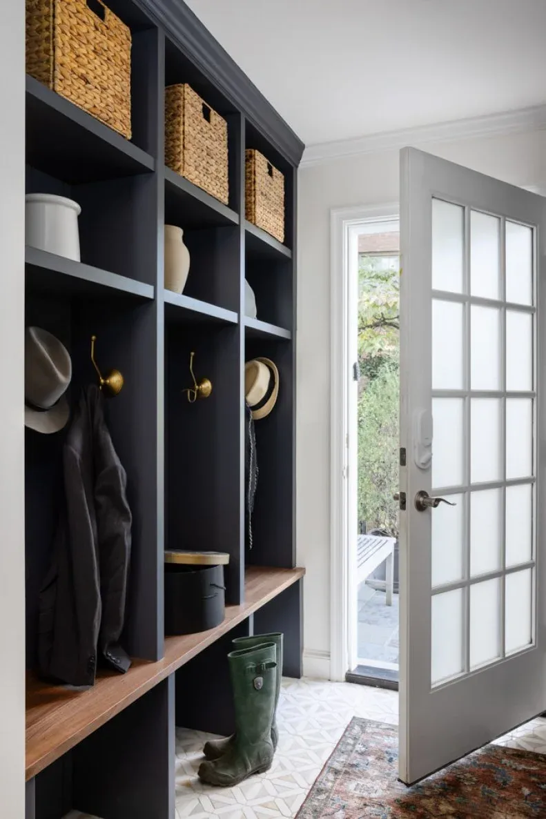 Mudroom with dark open coat cubbies, wooden bench surface, upper woven baskets, green boots and patterned rug.