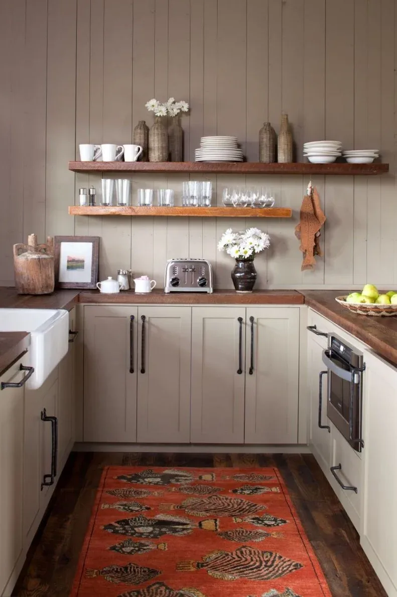 Taupe country kitchen with open wood shelves of dishes and glasses, apron-front sink, wood counter and a warm patterned rug