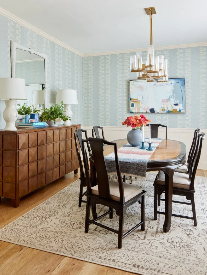 Wood dining sideboard with square relief fronts beneath a mirror and two white lamps beside an oval table