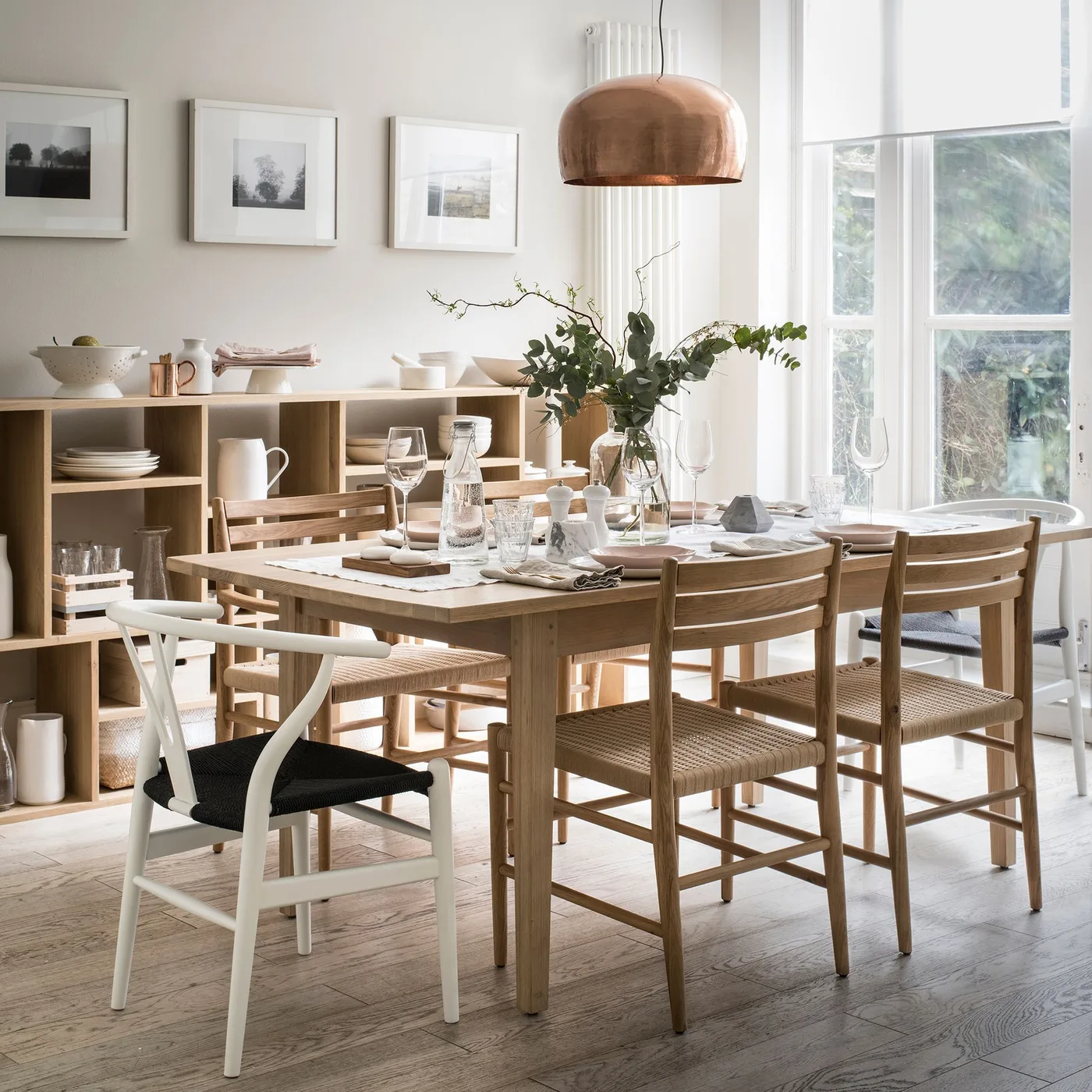 Pale dining room with a wooden table and chairs, low open wooden cubbies displaying dishes, framed pictures and a warm metallic pendant shade.