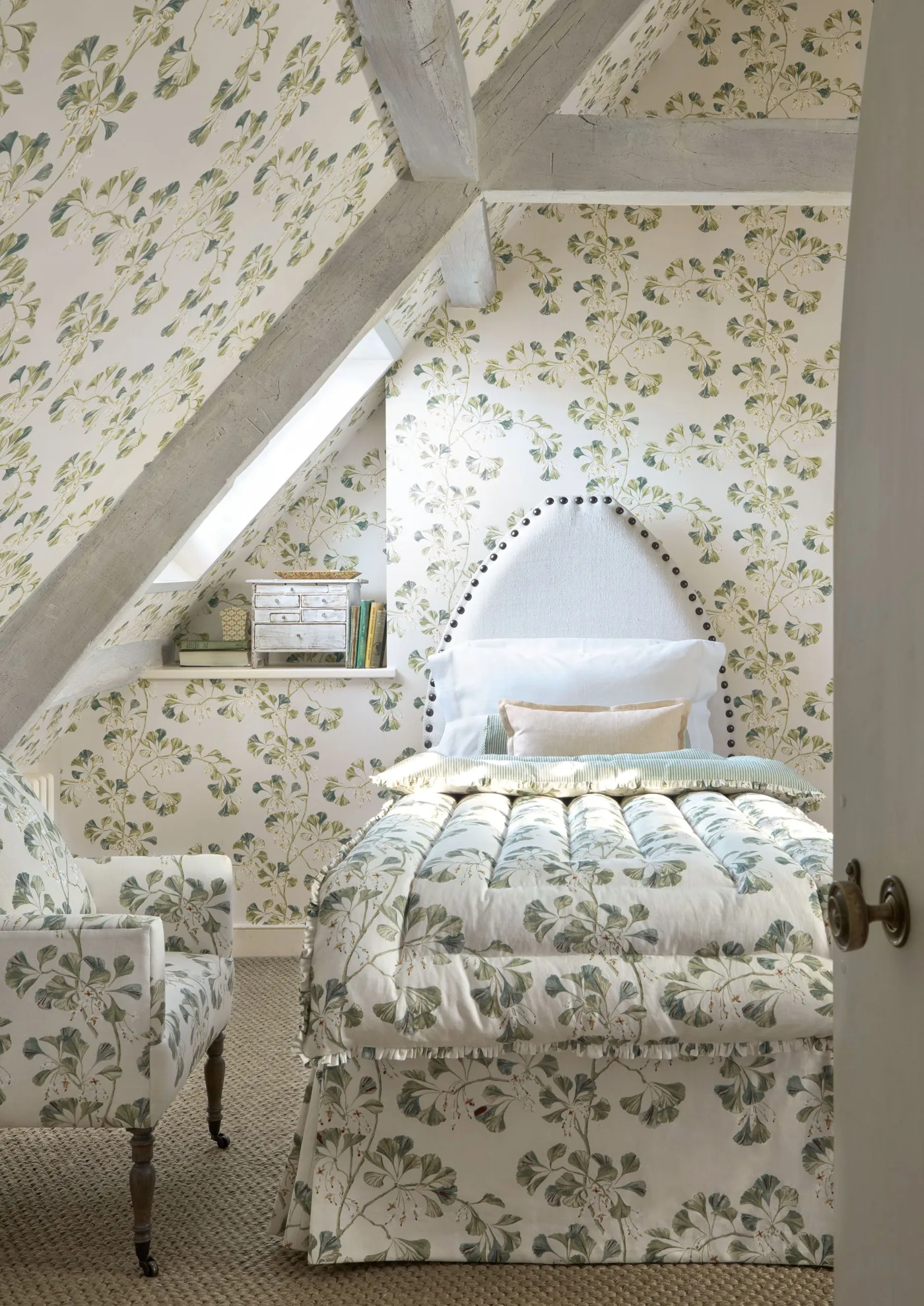 Attic bedroom with leafy wall and ceiling patterns, matching bedcover, a pale shaped headboard and a patterned chair.