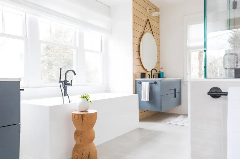 A white angular freestanding bath sits beside a small wood table and floating gray vanity beneath a round mirror on a warm paneled wall