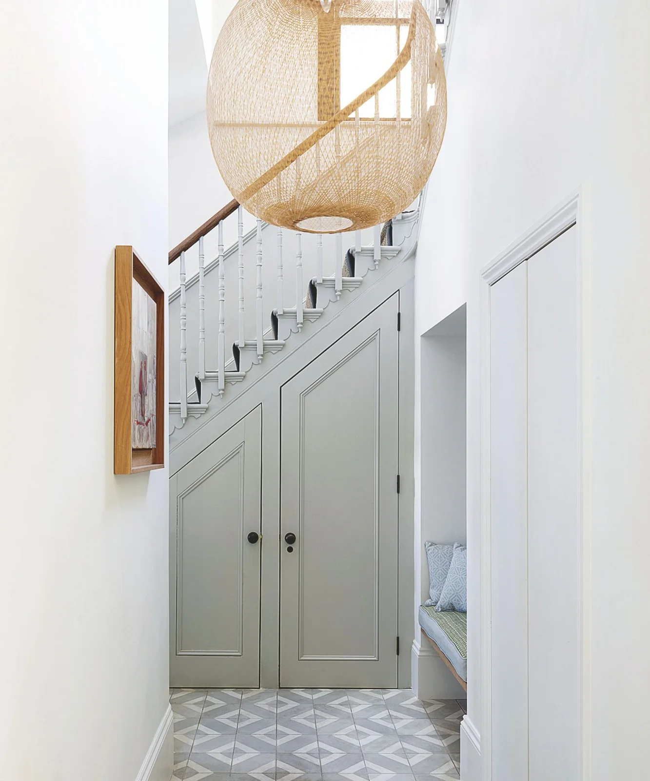 Pale green cupboard doors sit beneath a white staircase beside a recessed cushioned seat, geometric floor tiles and a large woven pendant