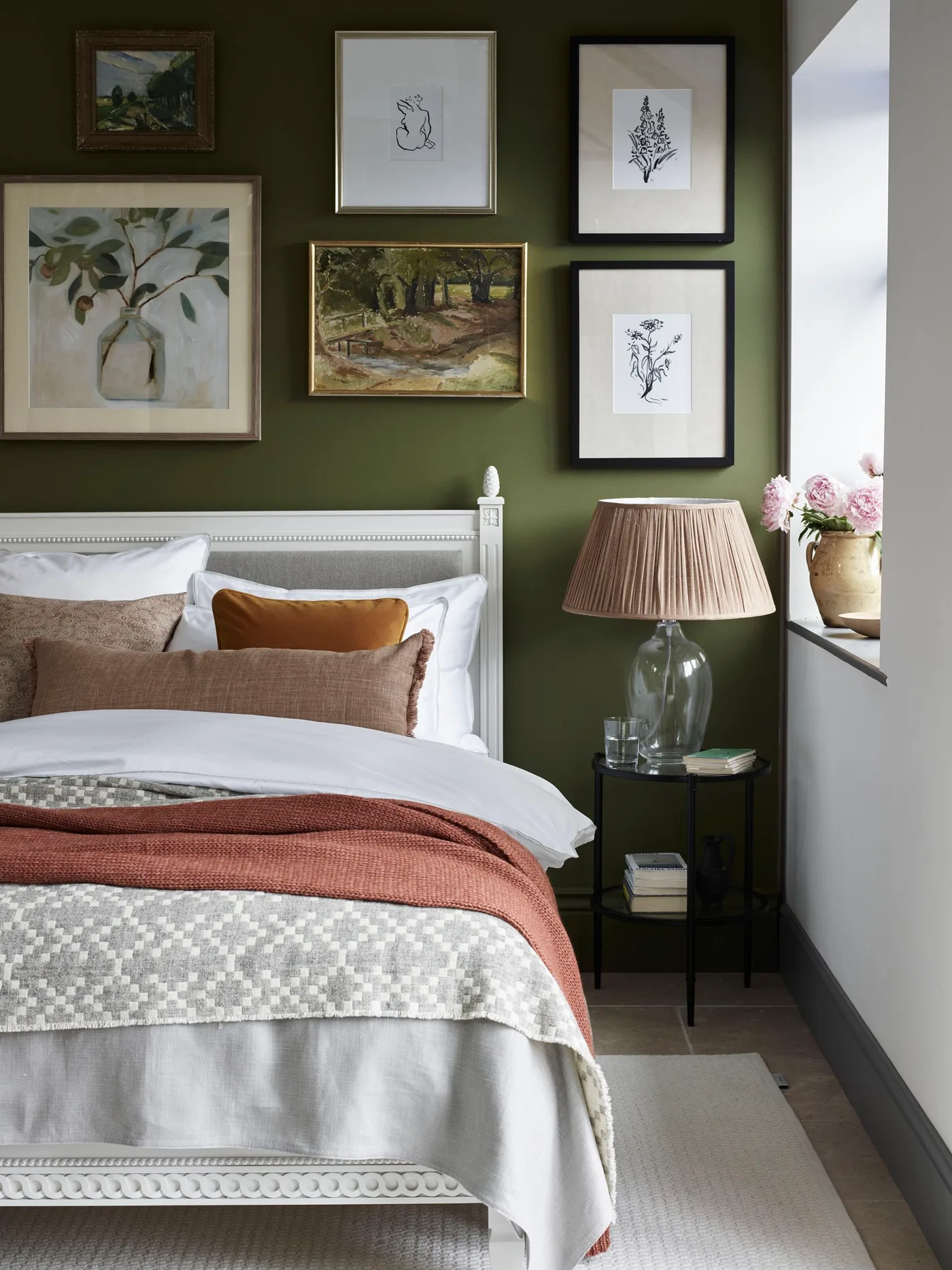 Olive green bedroom wall with a gallery of botanical and landscape pictures, pale painted bed, white bedding, rust throw, and glass-based lamp.