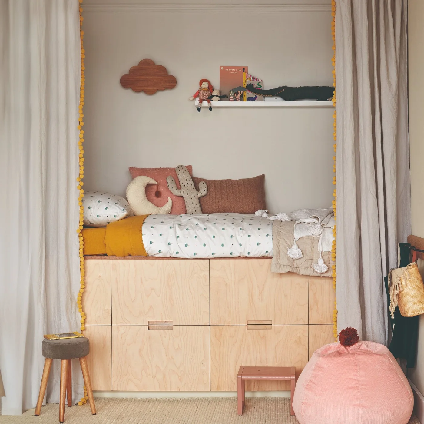 A sleeping nook with pale wood drawers below holds light bedding and pink cushions beside plain curtains, a pink floor cushion and a small step stool