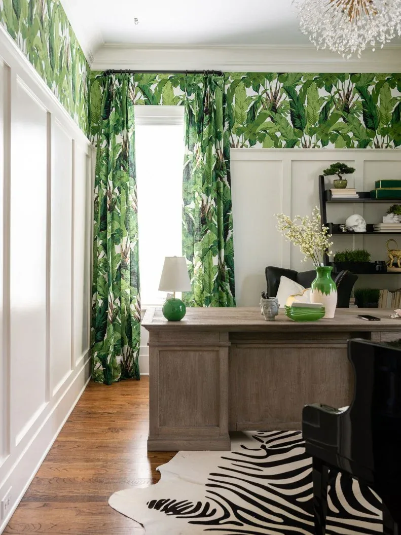 Green palm-patterned wallpaper and matching curtains surround a wooden desk above pale paneling beside dark office seating