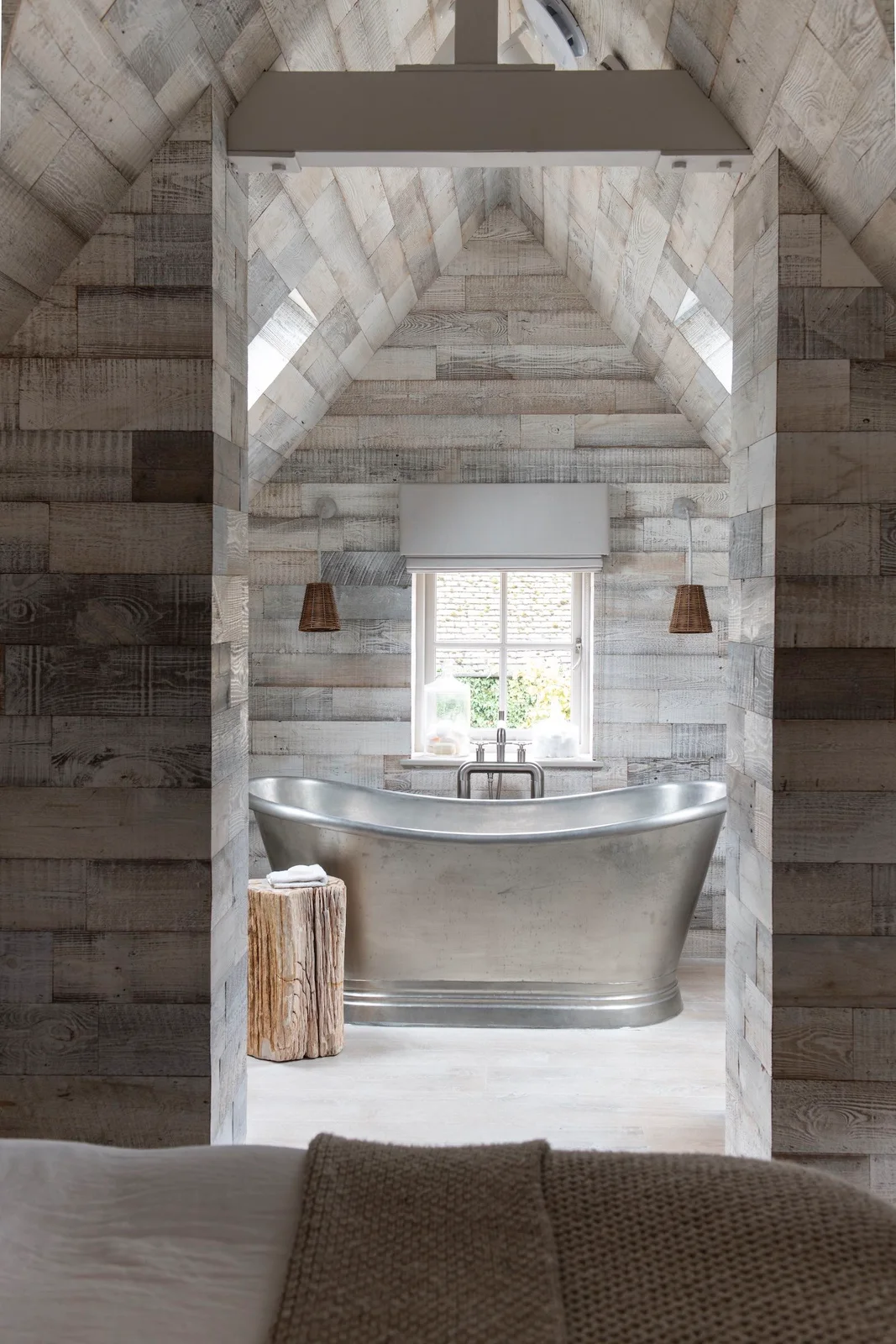 A smooth metallic-looking freestanding tub sits inside a pale wood-clad alcove beneath a window and two small woven hanging shades beside a wood stool