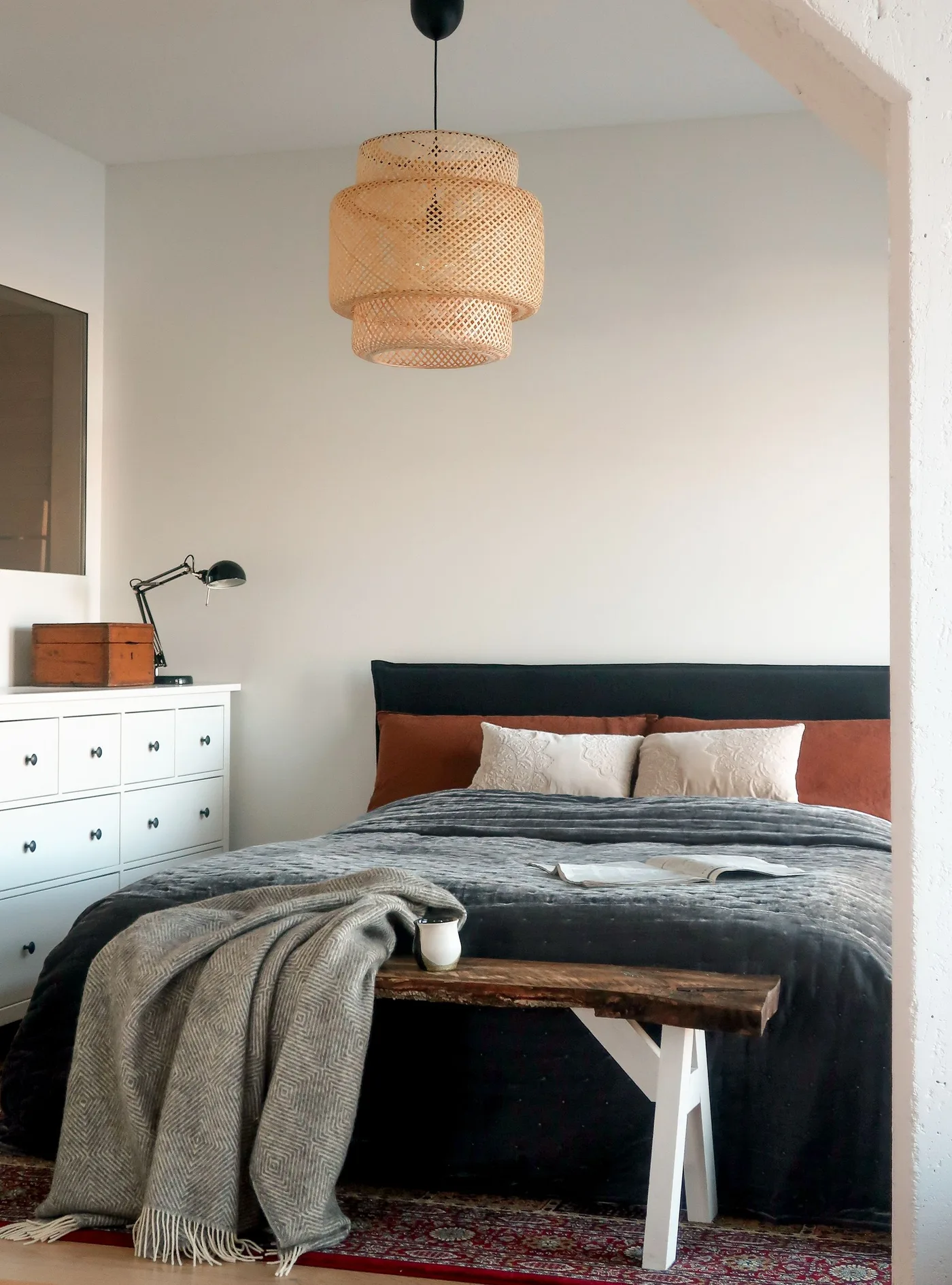 Woven pendant above a bedroom with rust pillows, charcoal bedding, a pale drawer chest and a wood bench