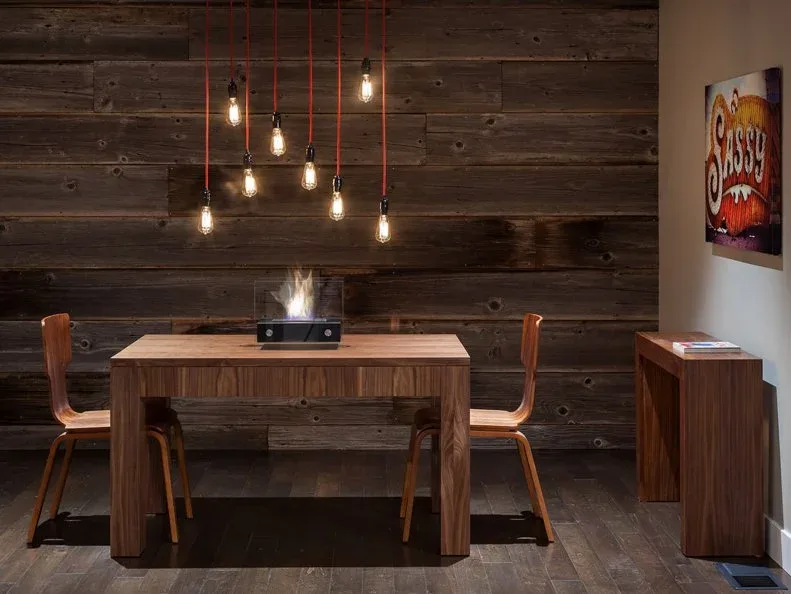 Two slim wooden dining chairs beside a thick wooden table and dark horizontal timber wall, with individual hanging lights overhead.