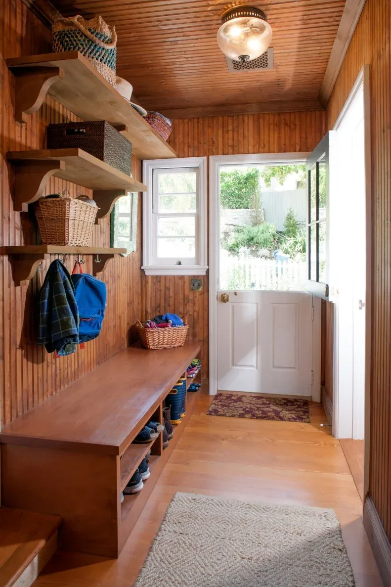 Wood-lined mudroom with long wood bench, lower shoe cubbies, upper shelves, wicker baskets and white Dutch door