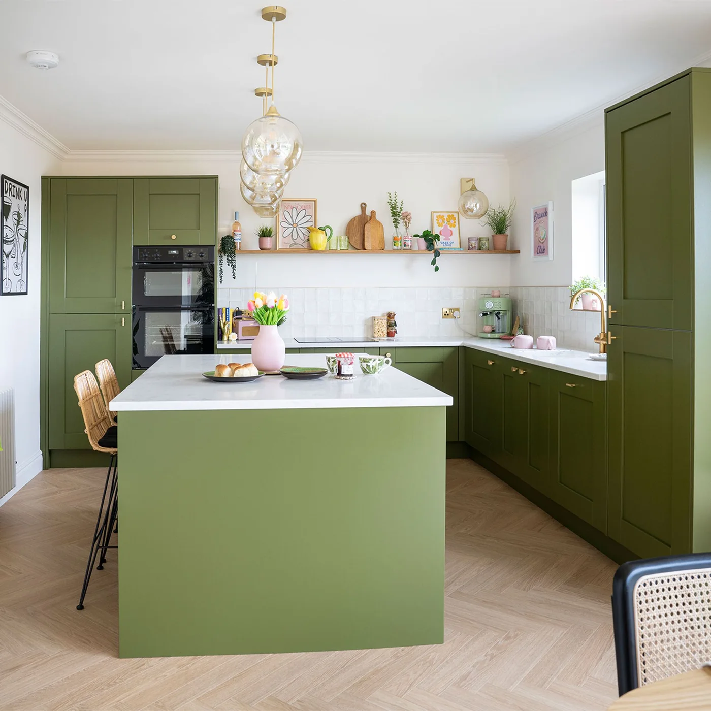 Olive green cabinets and an island sit beneath clear pendant lights beside white tiles and a narrow wooden shelf