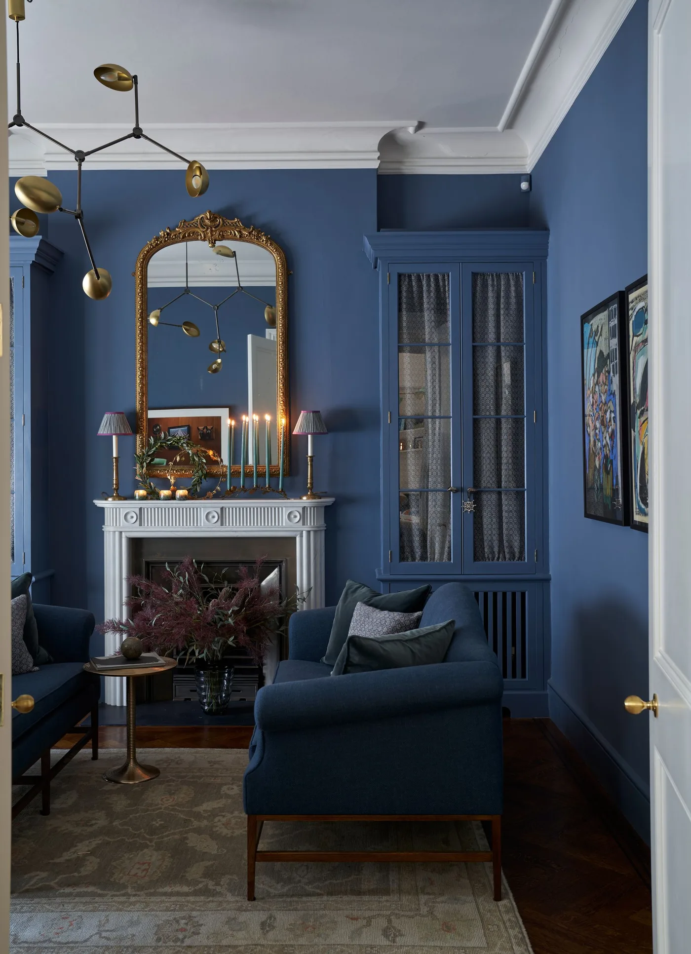 Blue sitting room with pale mantel, tall ornate gold-toned mirror, small lamps and slender candle holders