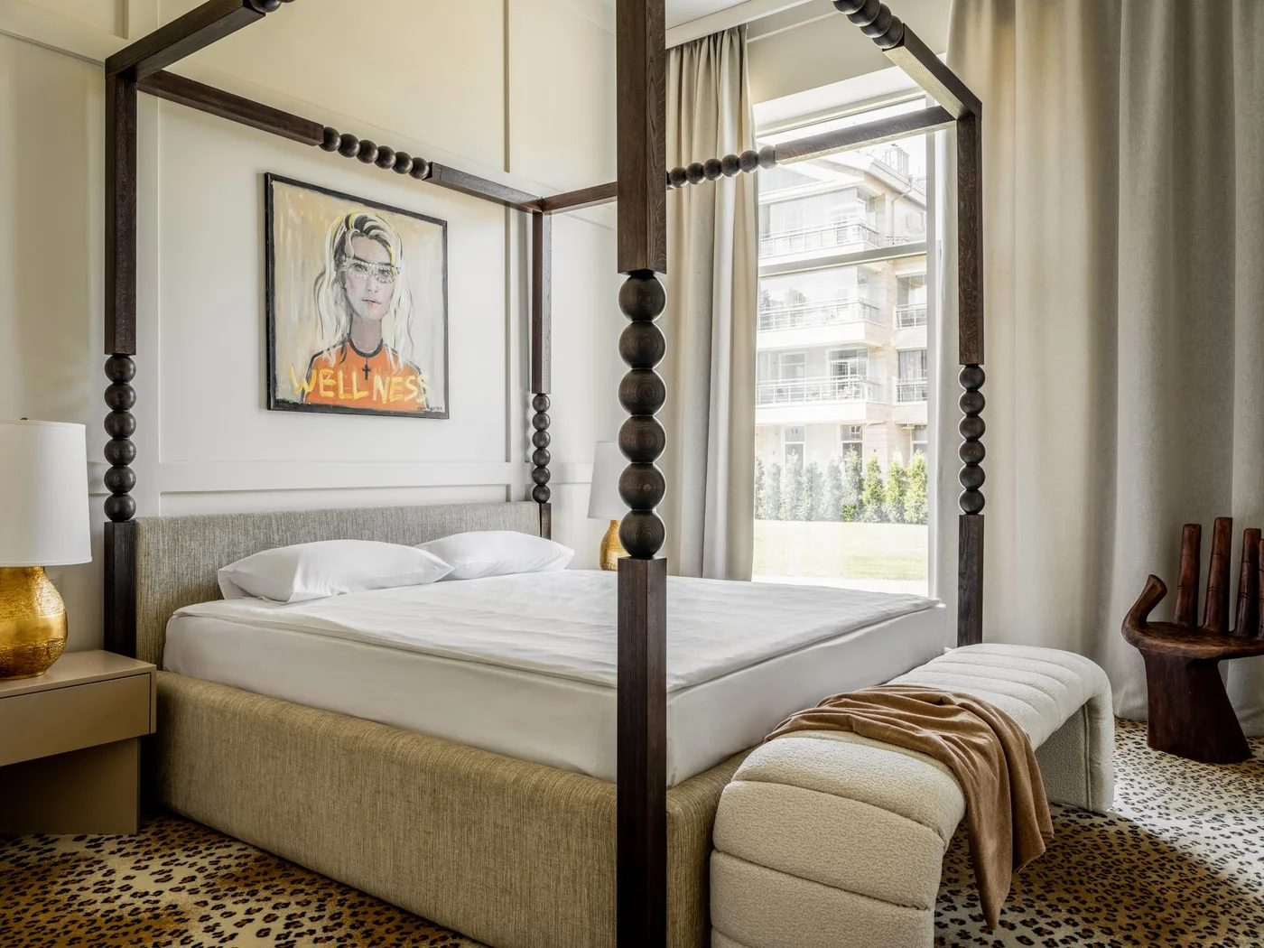 A dark four-poster bed with rounded details stands above a spotted gold-and-black rug beside a large graphic portrait and a curved pale bench