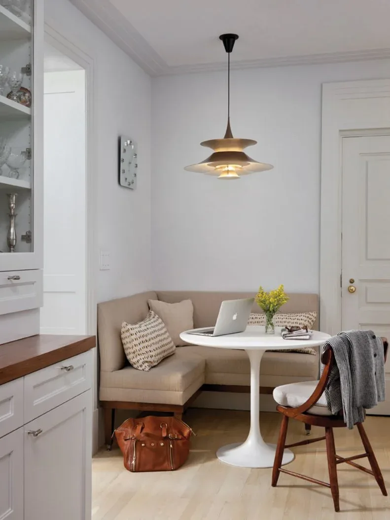 Pale beige corner banquette near white cabinetry, with a white round pedestal table, patterned pillows, a wooden chair, and a layered gold-colored pendant.