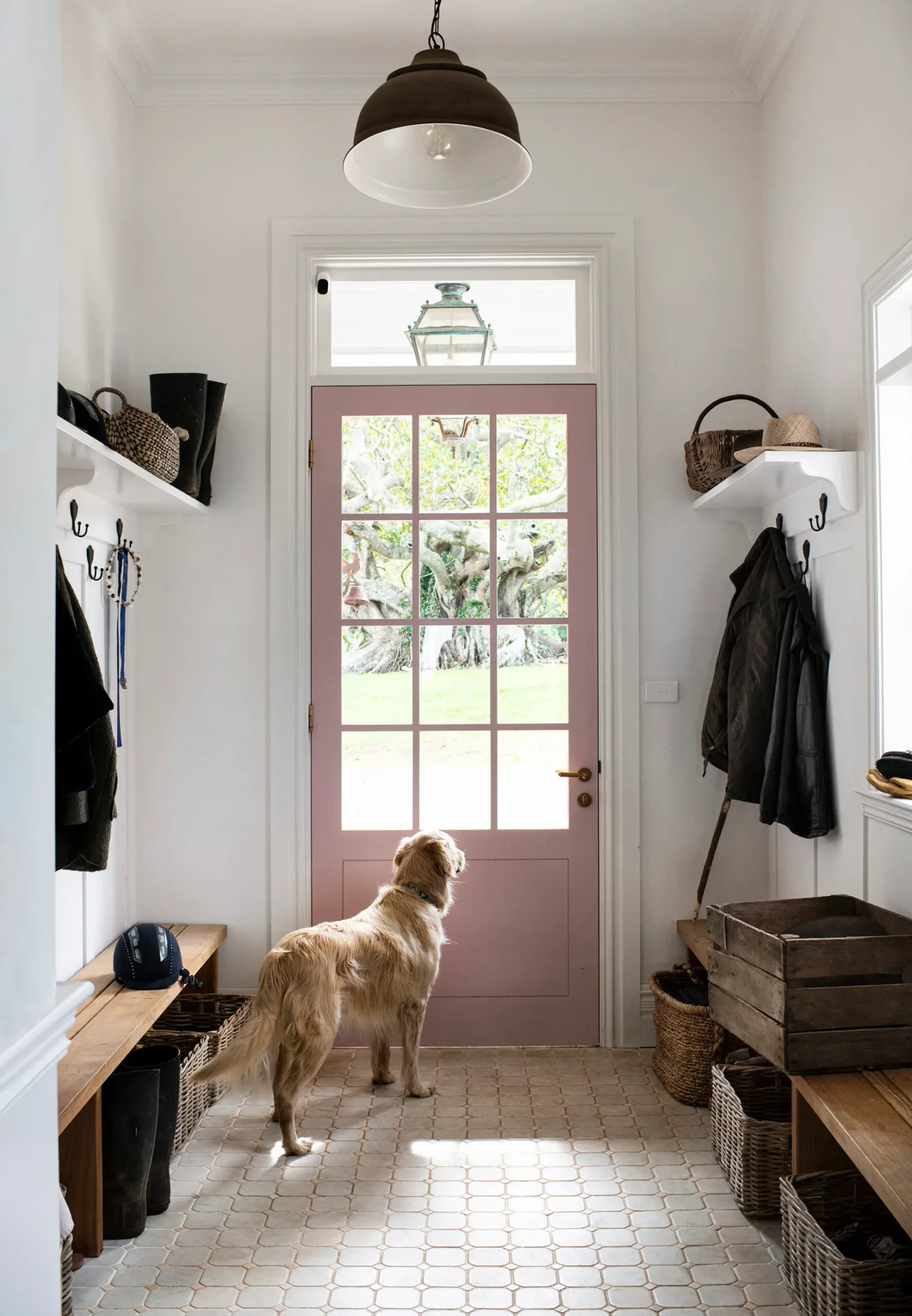 Country entryway with a pink glazed door, wooden benches, wall hooks, upper shelves, baskets and pale patterned floor tiles.