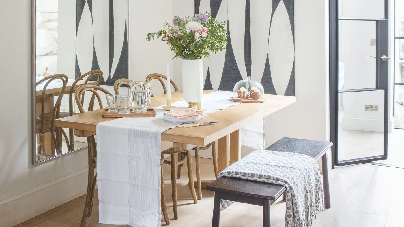 Pale wooden dining table with curved wooden chairs, a dark bench, patterned throw and a woven floor-lamp shade.