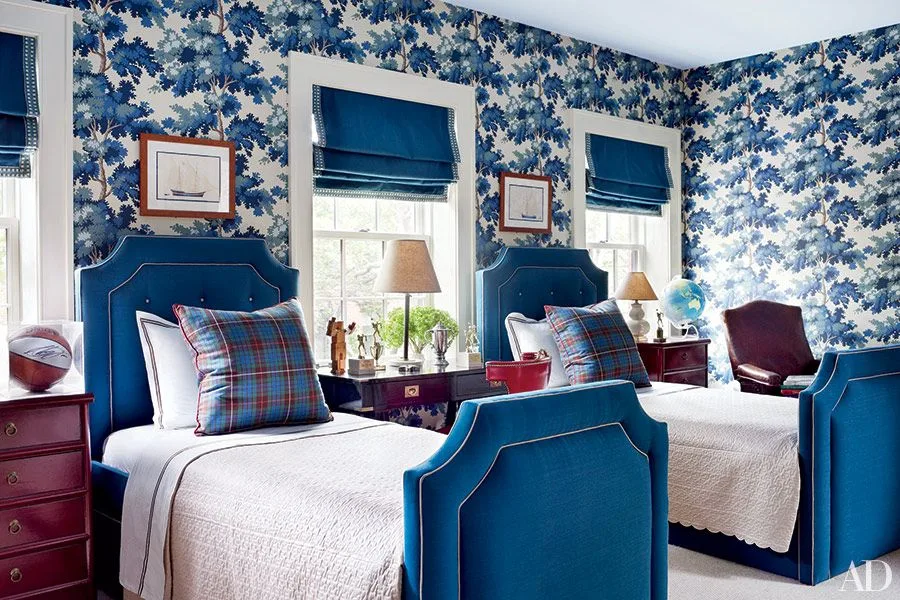 Two deep blue upholstered beds with white bedding, checked cushions, blue patterned wallpaper, and red bedside furniture.