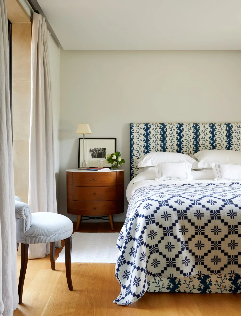 A bed with a blue patterned headboard and geometric blue cover stands beside a plain wood drawer chest, light curtains and a pale upholstered chair