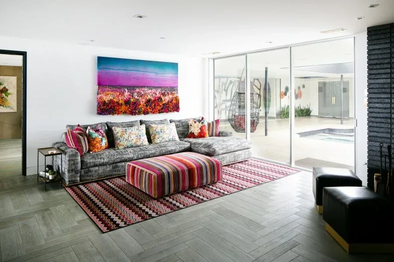 Living room with gray sectional, bright striped ottoman, red-and-cream patterned rug, mixed cushions, colorful landscape artwork and broad sliding glass doors.