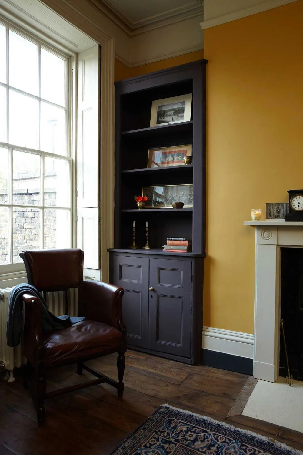 Ochre living room wall beside a tall dark bookcase, brown armchair and pale window trim