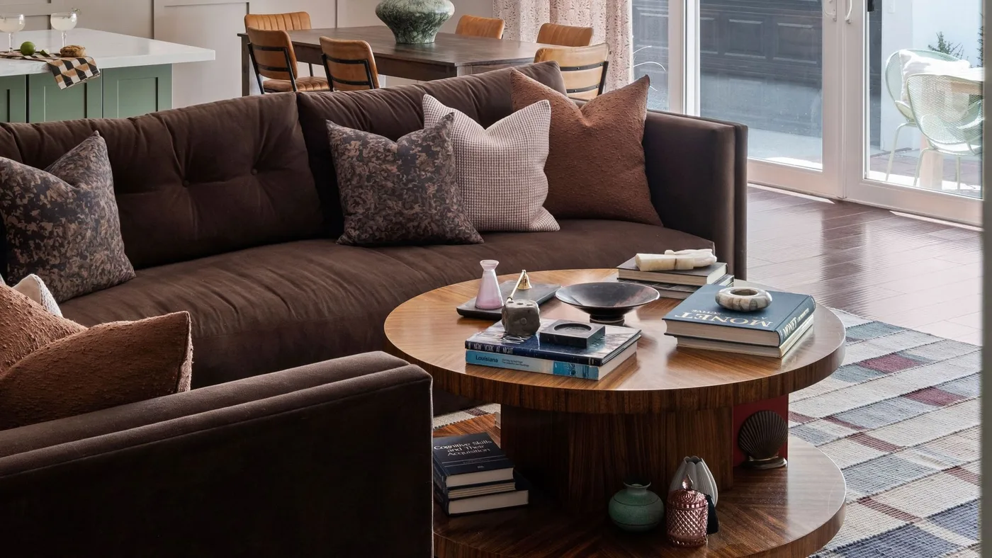 Brown sectional with rust, checked and floral pillows beside a round wooden table.