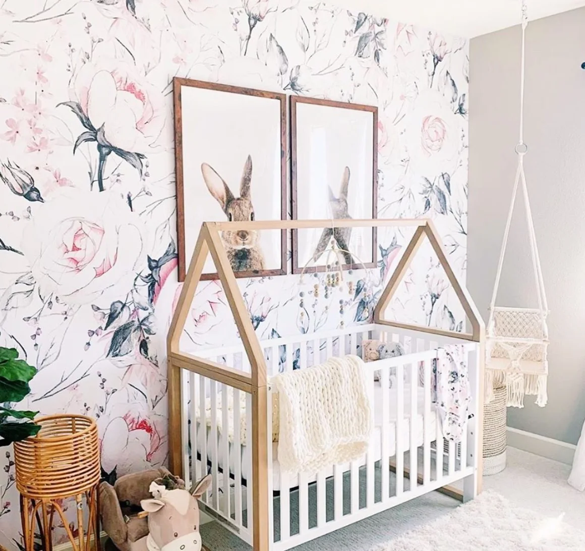 Pale pink floral nursery wallpaper forms a background for two large rabbit prints above a white crib with a wood-toned house-shaped frame