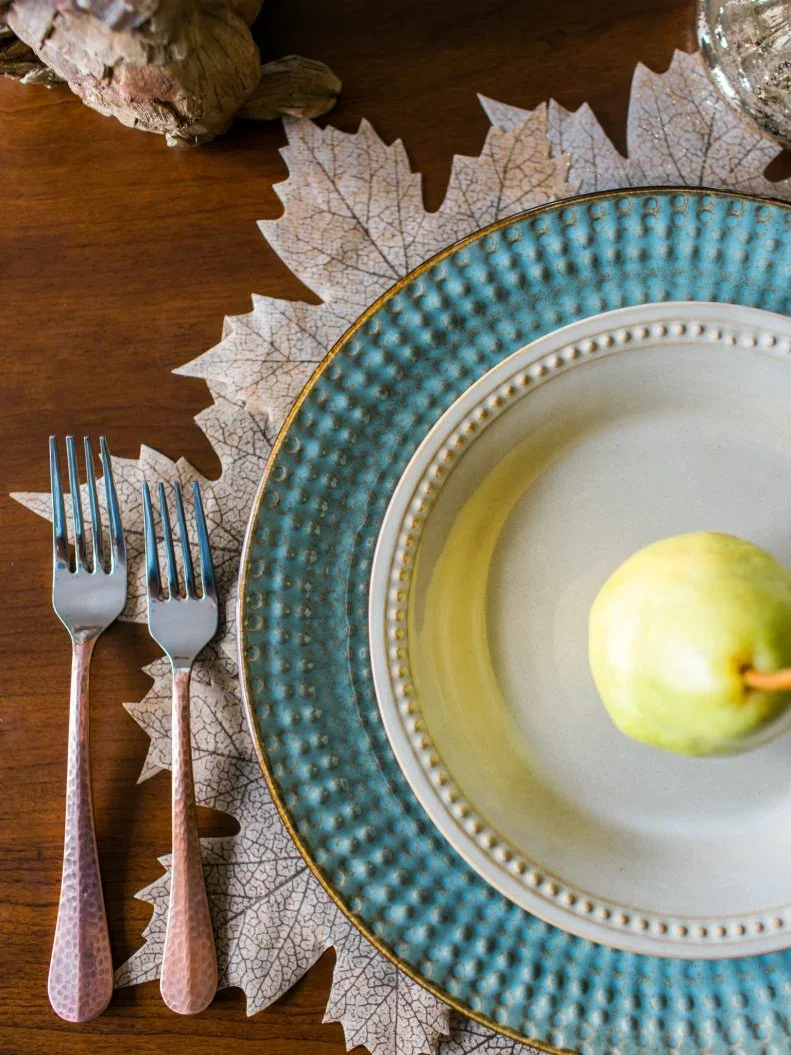 A green pear rests on a white plate above a textured turquoise charger and leaf-shaped mat beside warm-toned cutlery