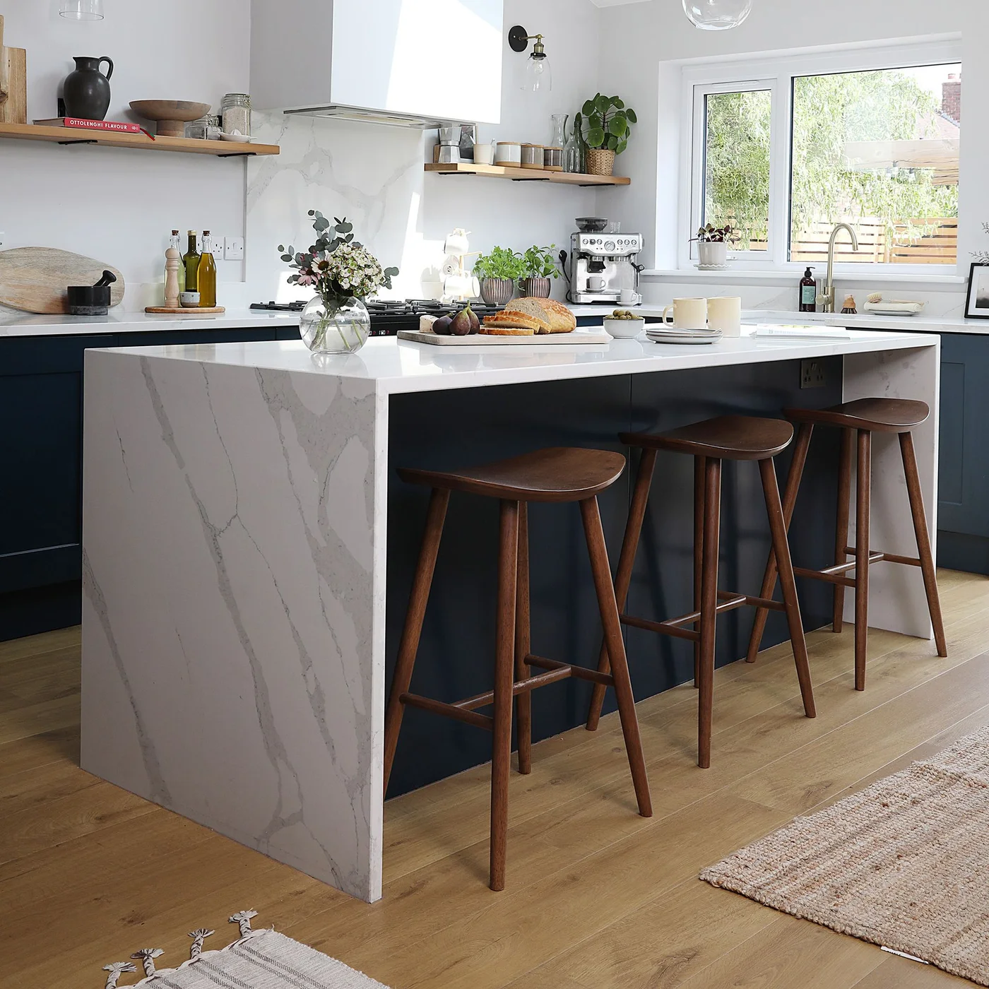 A dark kitchen island with a pale veined waterfall end holds three wood stools below lighter walls, a window and a simple extraction hood