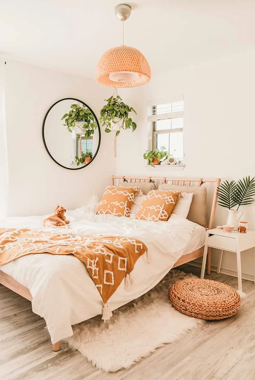 Pale bedroom with woven bed frame, orange patterned throw and pillows, hanging plants beside round mirror, woven ceiling light and round pouf.
