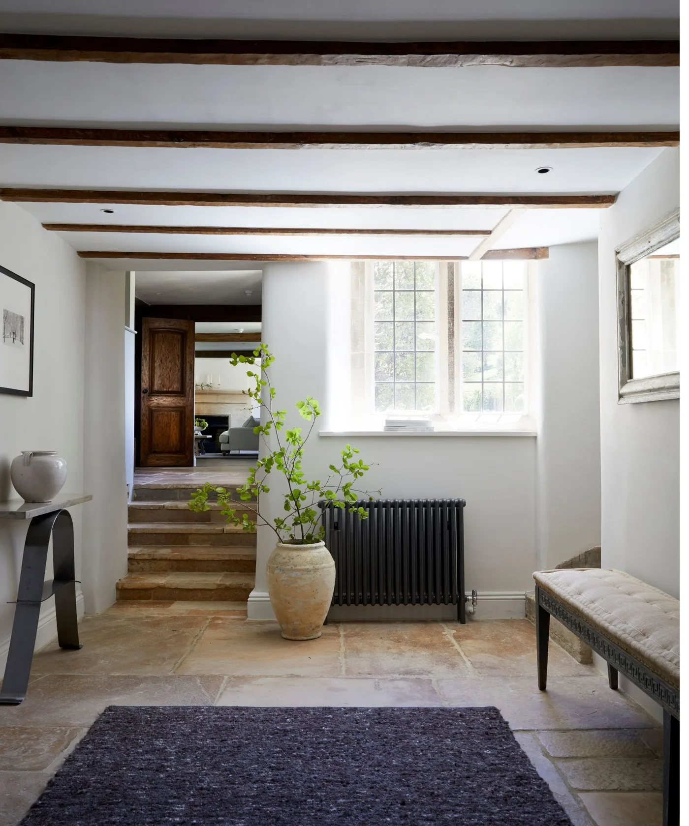 Dark textured rug on pale stone-looking floor beneath exposed beams, beside an upholstered bench and black radiator