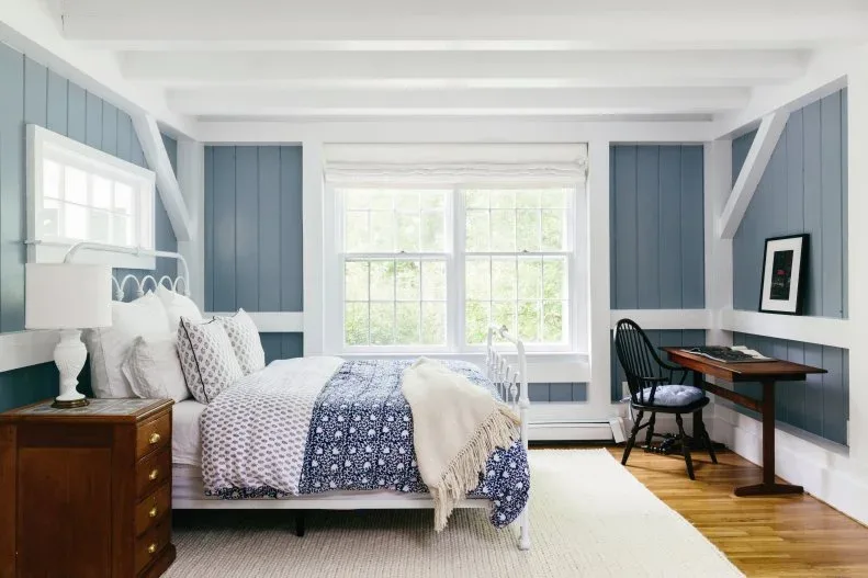 Blue paneled bedroom with white framing, metal bed, wood bedside chest and small writing table