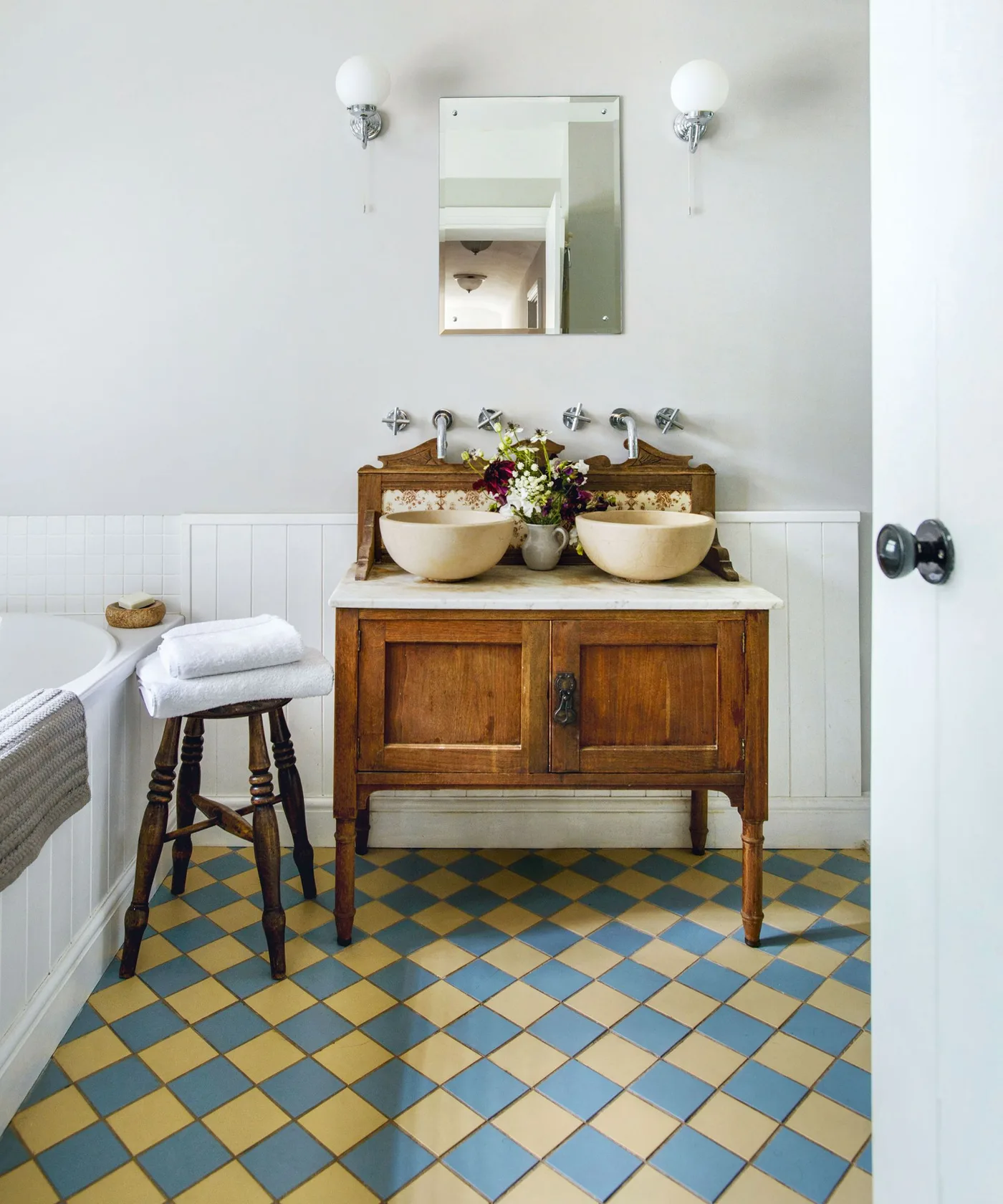 Wood vanity with two round basins above a blue-and-yellow checkerboard bathroom floor and white lower paneling.