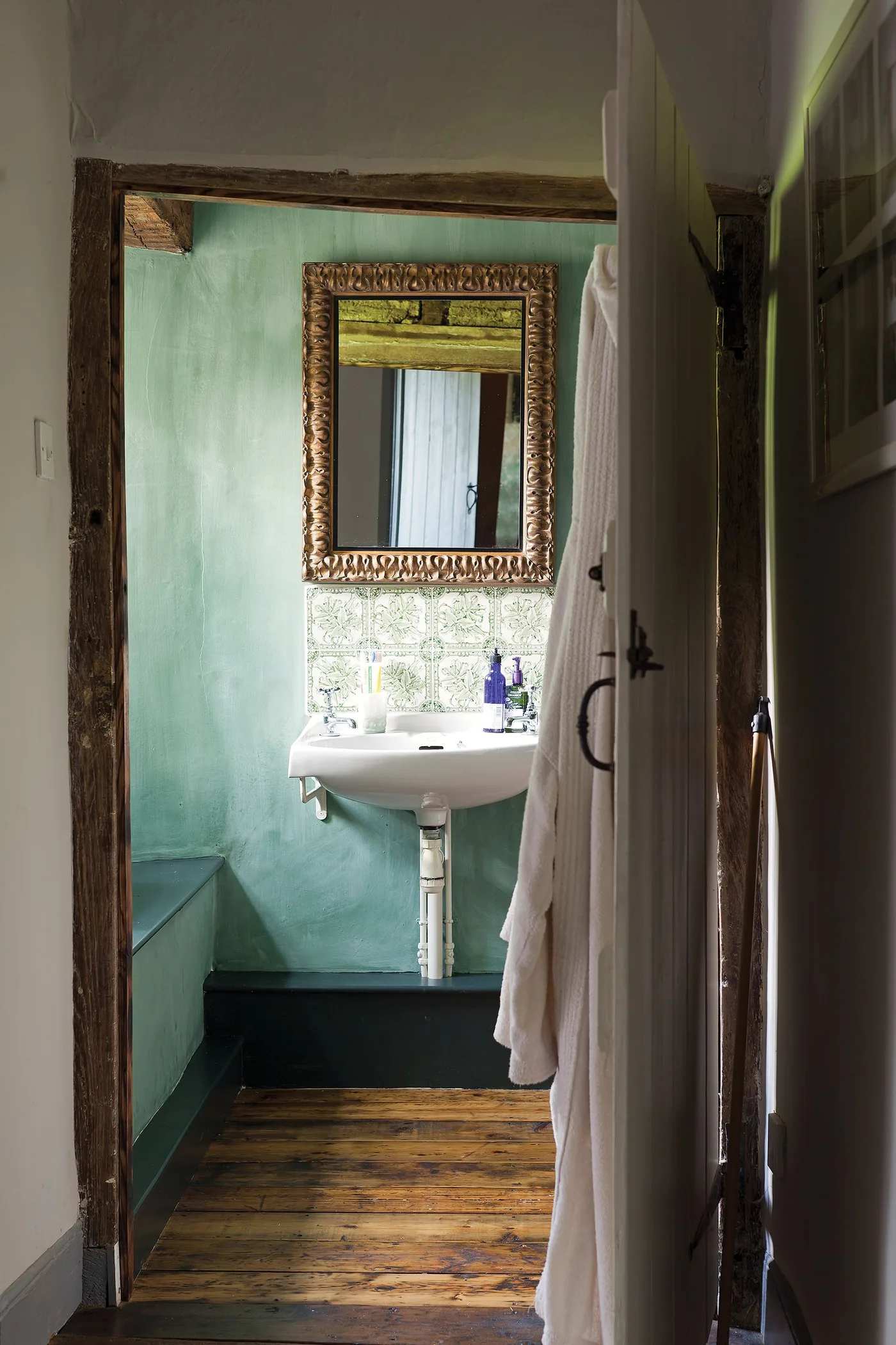 Compact bathroom with green-blue walls, a small white wall-mounted basin, ornate framed mirror, exposed beam and wooden floor.