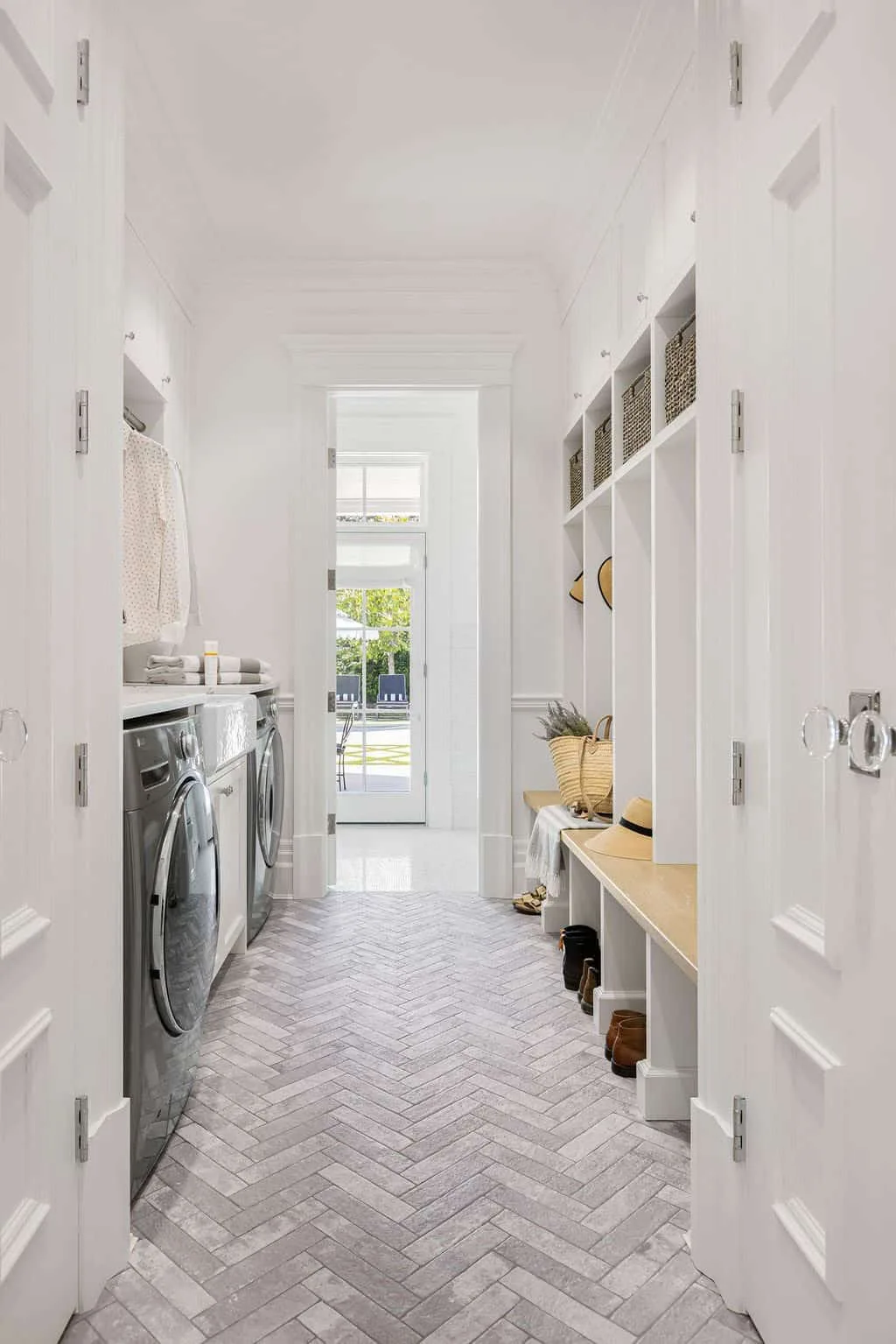 Narrow white laundry and mudroom passage with appliances opposite a bench and pale herringbone flooring.