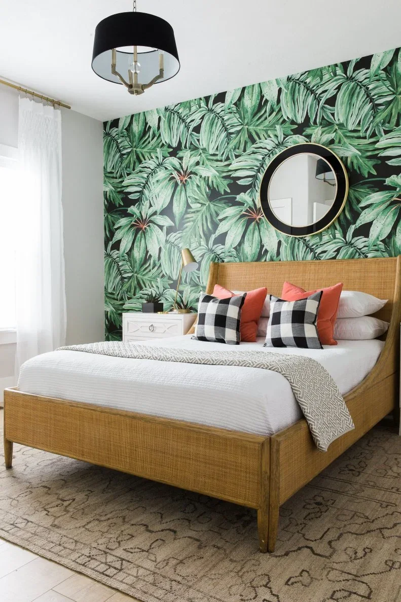 A green leaf wallpaper wall holds a round black mirror above a woven bed with white bedding, coral cushions and checked pillows