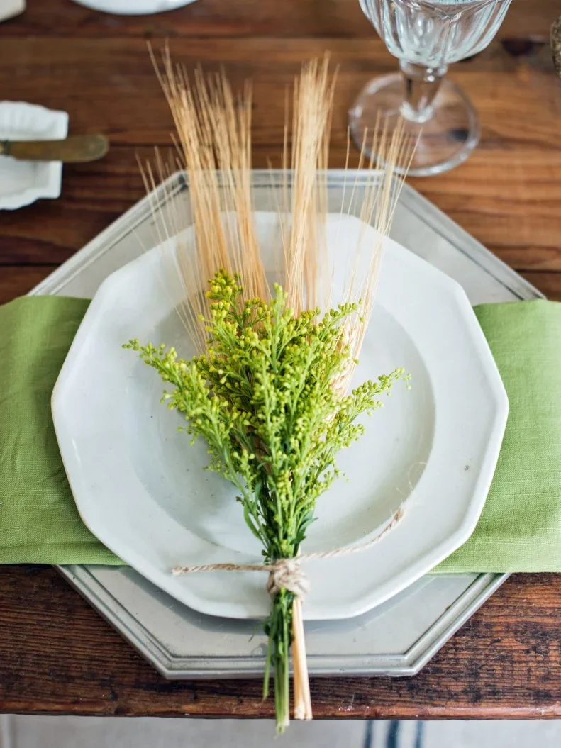 A tied bundle of pale wheat and bright green stems rests on white plates above a green napkin and gray charger